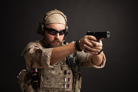 Brutal man in the military desert uniform and body armor standing in a fighting rack and aiming from his gun on a black background. Close-up of the gun barrel directed towards the camera