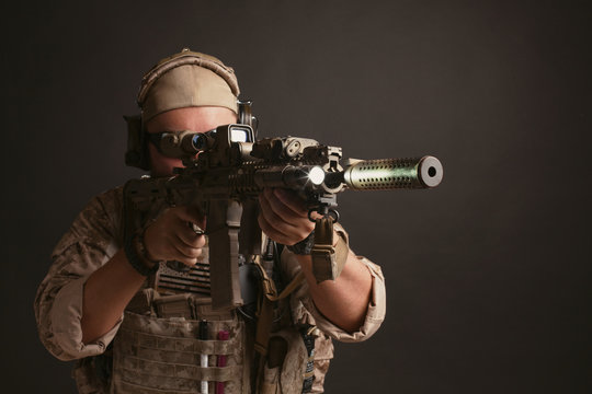 Brutal man in the military desert uniform and body armor stands in a fighting rack and holds his rifle on a black background. The bearded player in the airsoft safety glasses aiming a rifle