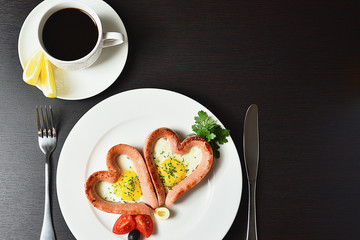 Scrambled eggs in the form of heart on a white plate with sausages, tomatoes, greens and coffee with lemon.