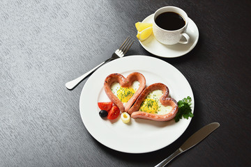 Scrambled eggs in the form of heart on a white plate with sausages, tomatoes, greens and coffee with lemon.