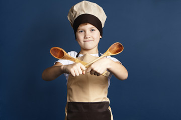 Happy confident 10 year old male child in chef headwear and apron holding two wooden spoons in...