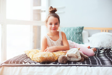 happy child girl wake up in the early morning in her room, lying on comfortable bed in cozy homely interior