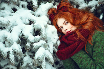 Close up portrait of beautiful red haired girl with creative hairstyle decorated with knitted flowers leaning her head against snow covered fir branch and warming her hands under scarf