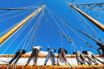 Foto auf Acrylglas Segeln Die Besatzung lehnt sich über die Seite eines großen Segelboots, um das schwere Segel auf einem Großsegler auf See zu heben. Es braucht sechs starke Leute, um das Segeltuch zu hissen. Thema für Teamarbeit, Zusammenarbeit