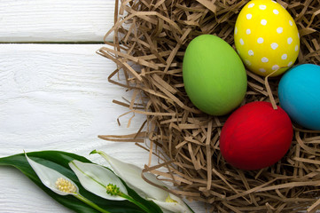 Colored easter eggs in nest with flower top view on white wooden background