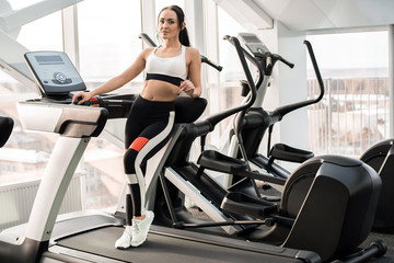 Fototapeta na wymiar Full length portrait of fit young woman posing standing on treadmill in modern gym, copy space