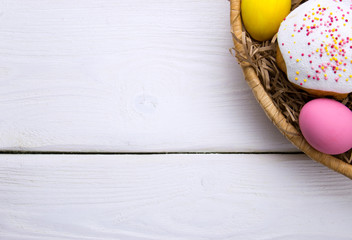 Colored easter eggs and little easter cake in nest beasket on white wooden background top white