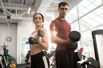 Waist up portrait of fit sportive couple posing with dumbbells looking at camera in modern gym