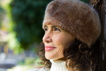 Bright close up portrait of dazzling mature woman wearing hairy fur beret hat and leaning on tree in the park. Middle aged lovely smiling lady portrait outdoors