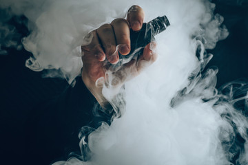 Man hand shows vape device at cloud of vapor background. Vape concept