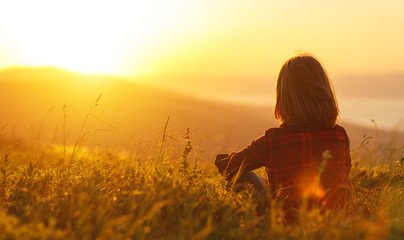 Woman sits with her back in the field and look sunset in the mountains