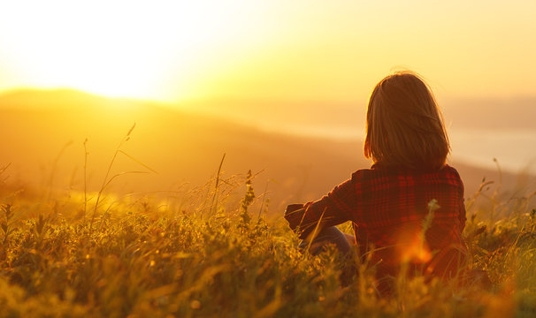 Woman sits with her back in the field and look sunset in the mountains