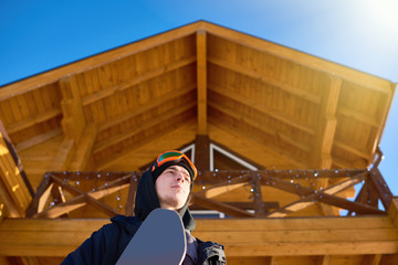 Low angle portrait of handsome young snowboarder against wooden chalet at modern ski resort enjoying sunny winter, copy space