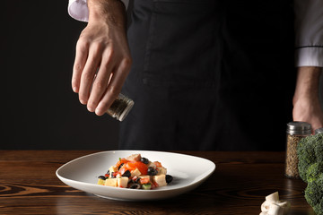 Male chef in uniform adding spices to tasty dish on table, closeup