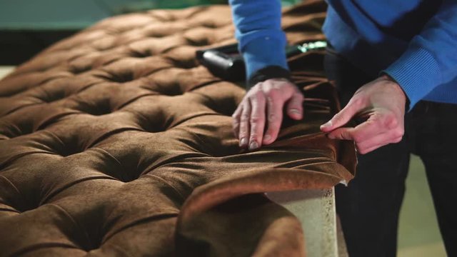 Cabinet maker is wrapping a sofa with a cloth. He is tightening fabric and fixing it with industrial stapling machine in a workshop of factory