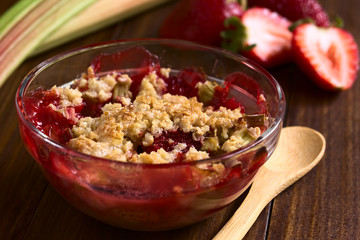 Baked strawberry and rhubarb crumble in glass bowl with fresh raw ingredients in the back, photographed on dark wood with natural light (Selective Focus, Focus one third into the bowl)