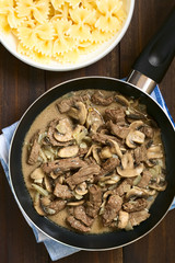 Beef Stroganoff in frying pan, a dish made of pieces of beef, mushroom and onion in cream sauce, with pasta on the side, photographed overhead with natural light