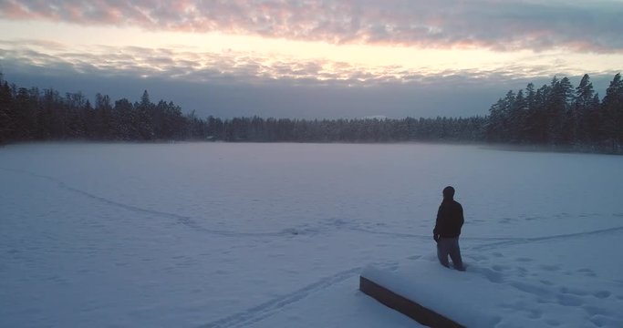 Man standing at a dock, Cinema 4k aerial sideway view of a hiker on a warf, at a frozen smokey, winter lake, on a cold winter day, in Uusimaa, Finland