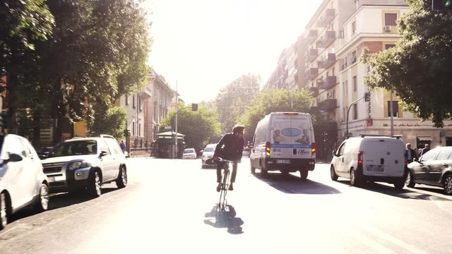 Young hipster man riding bike in traffic on a road with trees in Rome city centre wearing sunglasses on sunny day slow motion camera car steadycam