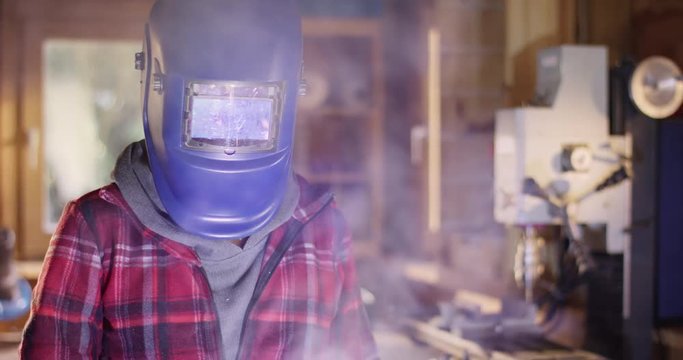 Young attractive woman welds metal in a workshop and reveals her beautiful face