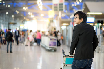 Young asian man with his luggage on airport trolley waiting for check in at airline counter in the international airport terminal, business travel concept