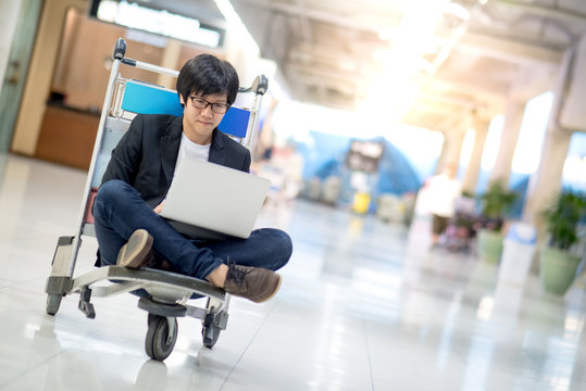 Young asian man working on airport trolley with his laptop computer during waiting for a connecting flight, freelance lifestyle and digital nomad concepts