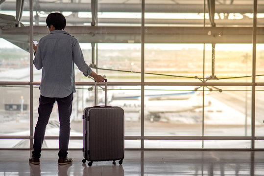 Young Asian man standing with suitcase luggage and holding smartphone in the international airport terminal, travel lifestyle concept