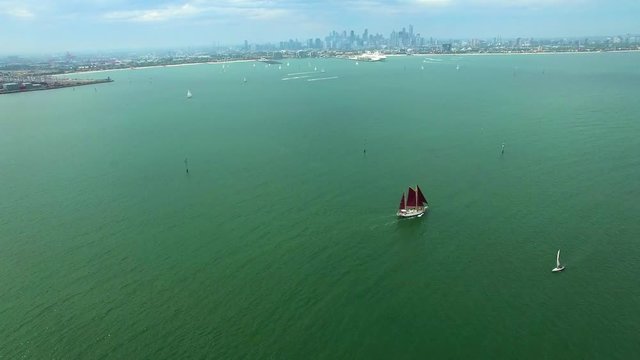 Static aerial shot of beautiful sailboat with dark red sails sailing across ocean bay with modern city skyscrapers skyline in the distance