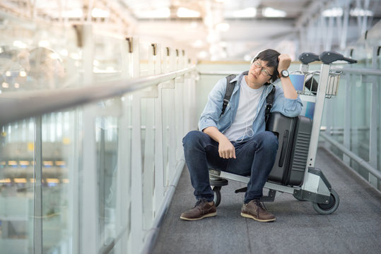 Young Asian man feeling exhausted sitting on airport trolley with his suitcase luggage in the international airport terminal, flight problem and travel insurance concepts