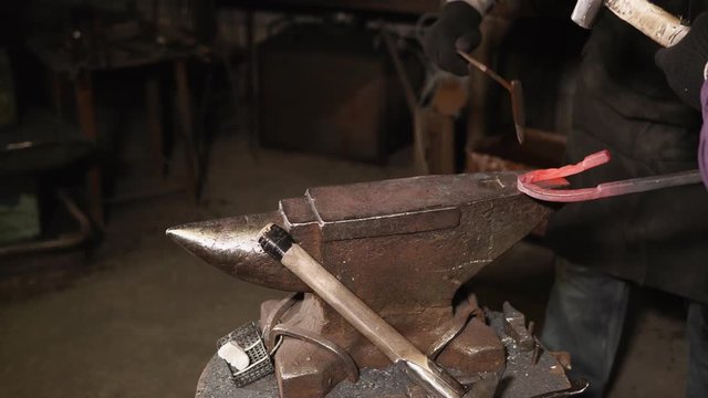 Master smith and his apprentice are processing metal detail in shop. One is holding horseshoe and small mallet, second is hitting over anvil