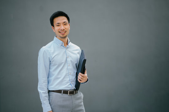 Portrait Of Happy Smiling Young Businessman, Standing On  A Plain Gray Background While Holding His Leather Folio. He Is Well-dressed In Business Casual And Looks Relaxed.