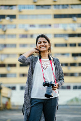 An  image of a very attractive Indian woman smiling ah she fix her hair with poise. She is wearing a casual outfit and holding her camera for her day tour.