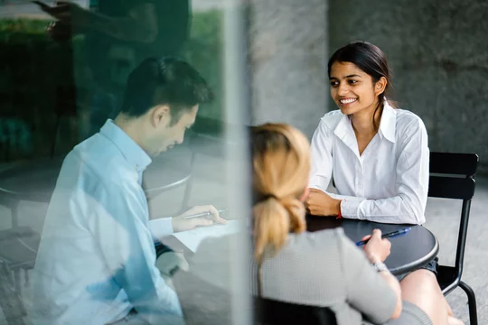 A young and attractive Indian Asian woman is interviewing for a job. She is dressed professionally in a white shirt and is sitting and talking to her interviewers. She is confident and relaxed