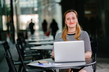 Portrait of a young but confident and mature Russian woman graciously smiling while working on her laptop. She sits at a table and wears a professional gray clothes.