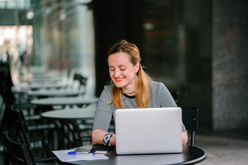 A professional, smooth and well - placed portrait of a Russian woman sitting in her office while working with some documents on her laptop. She's smiling as she's focused on completing today's task.