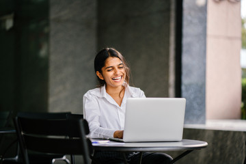 A portrait of a young job-seeker sitting at a table while looking for a jjob using her laptop. She is smiling as she types on her laptop and she is wearing a very casual outfit.