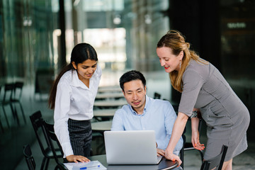 A diverse multi-racial team of three colleagues businesspeople have a relaxed discussion around a laptop notebook computer in an office or coworking space. They are all smiling and talking animatedly.