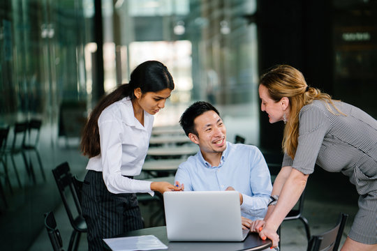 A diverse multi-racial team of three colleagues businesspeople have a relaxed discussion around a laptop notebook computer in an office or coworking space. They are all smiling and talking animatedly.