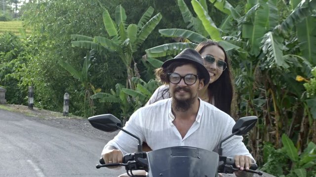 Beautiful Asian woman smiling and embracing her boyfriend while riding together on scooter on tropical road