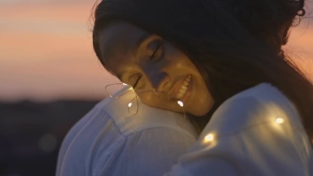 Closeup shot of happy young couple tangled in fairy lights hugging and smiling on rooftop date at sunset