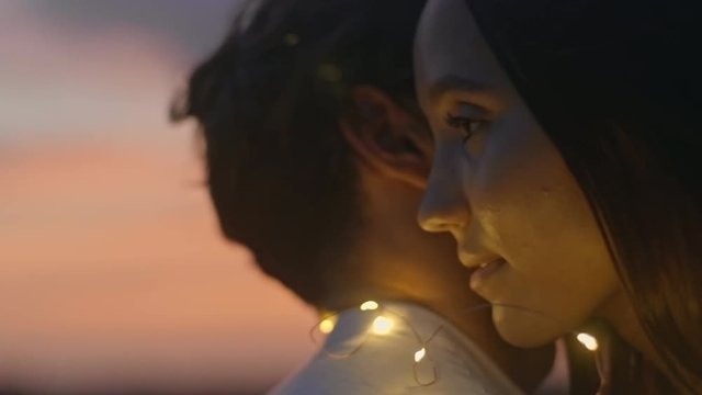 Young beautiful woman embracing boyfriend and whispering love confession in his ear. Romantic couple tangled in fairy lights standing on rooftop and enjoying romantic date