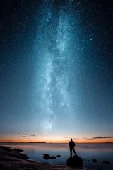 Silhouette eines Mannes, der allein auf einem Felsen am Meer steht und den Sonnenuntergang und die Sterne der Milchstraße betrachtet © Jamo Images