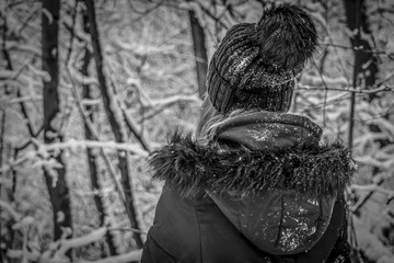 Girl from behind in a snowy forest, black and white,  Teenager girl in snowy  black coat, blurred forest in background, winter theme.