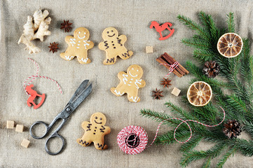 Gingerbread in the form of little men on a canvas background. The Christmas composition is supplemented with the root of ginger, anise, cinnamon. In the frame scissors and thread. View from above.