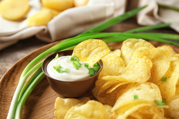 Crispy potato chips with green onion and sour cream on wooden plate, closeup