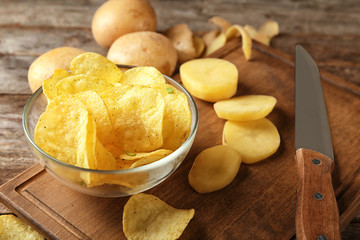 Bowl with crispy chips and raw potato on wooden table