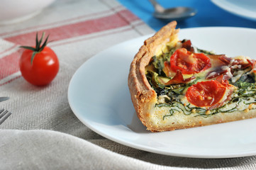 Portion of French pie quiche on a white plate. Filling with spinach, bacon, cheese, cherry tomato. In the frame, a cherry tomato. The background is draped with a napkin. Close-up. Macro photography.