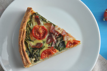 A piece of classic French pie quiche on a white plate. The composition of the filling is spinach, bacon, cheese, cherry tomatoes. View from above. Close-up. Macro photography.