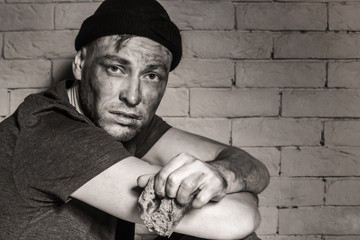 Hungry poor man with piece of bread near brick wall