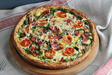 On a wooden dish, a pie quiche with bacon and spinach. When cooking, cheese and tomatoes are used...